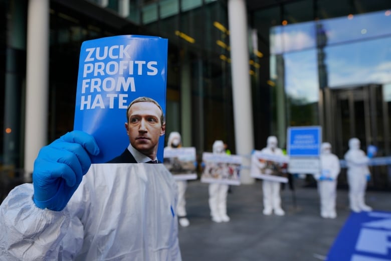 A man wearing gloves holds up a sign with Mark Zuckerberg's face on it.