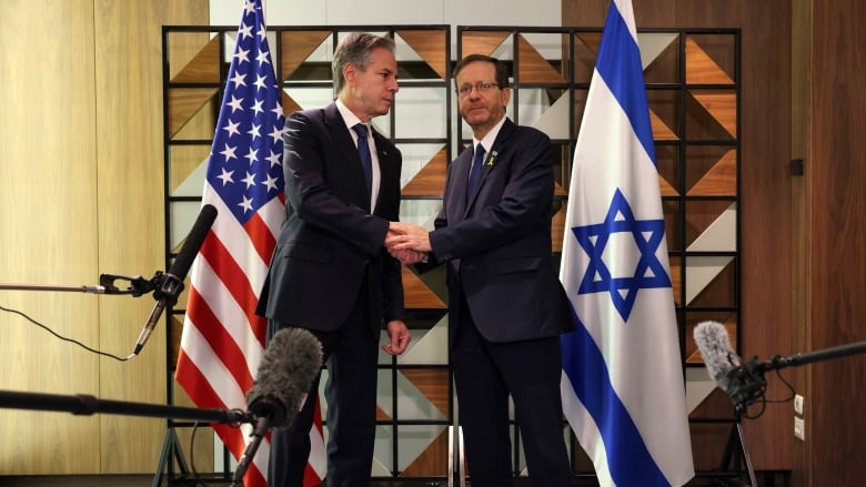 Two people shake hands in front of flags as microphones and boom mics are seen in front of them.