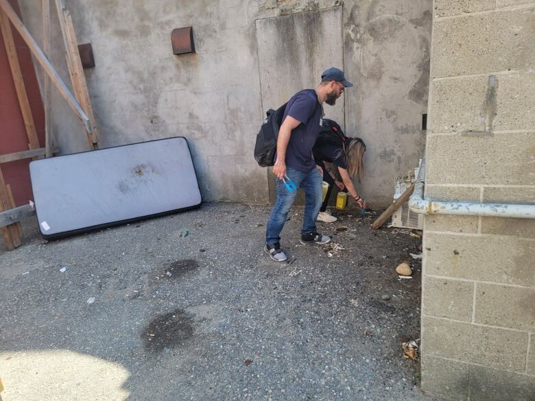 Two people pick up needles in an alleyway using tongs.