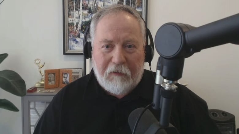 A man with a greyish beard and short hair wears headphones as he sits in front of a microphone.