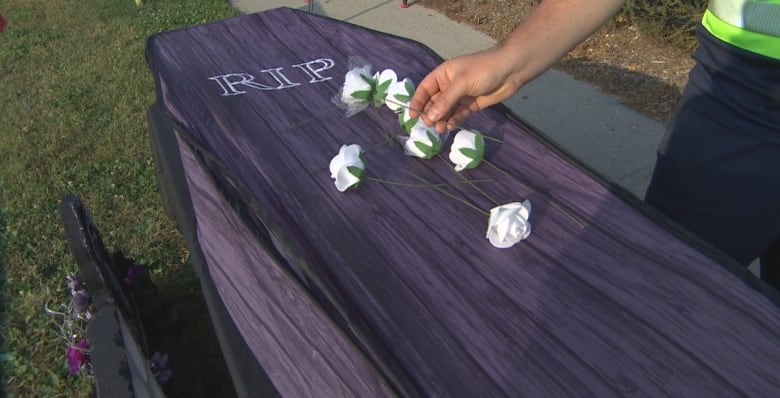 A person in a reflective vest lays a white rose onto a cardboard coffin. 