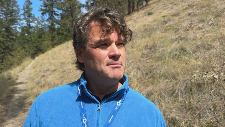 A man in a blue jacket is photographed with a few pine trees on a slope in the background.