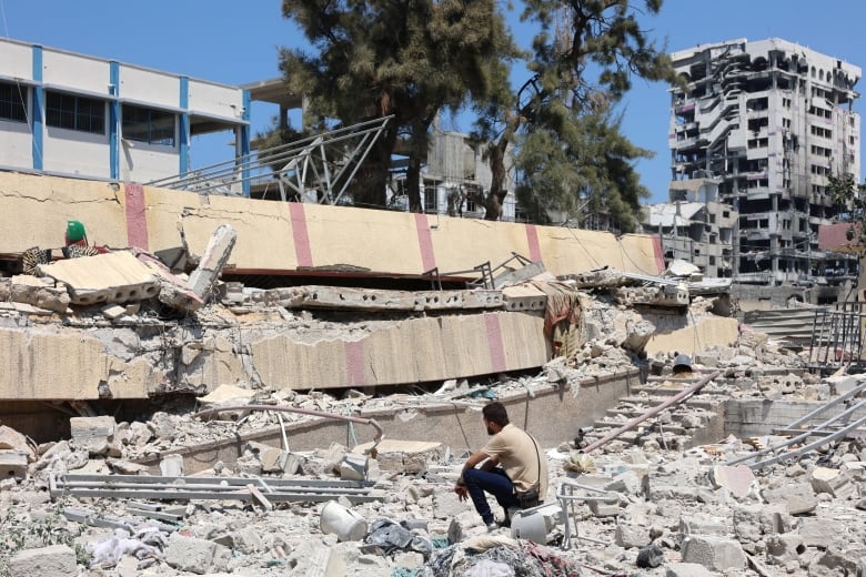 A man sits near concrete debris and a heavily damaged low-rise building. In the distance, a damaged 10-storey building is shown.