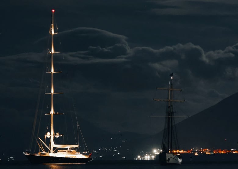 A nighttime view of a yacht with a tall mast, with lights on it, moored on the water with the lights of a city in the background. 