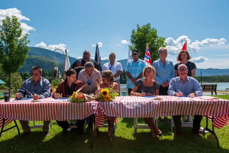 A long table outdoors with a number of people seated along its length with pens in had with the B.C. flag and various other flags behind them.