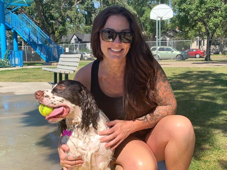 A woman poses with a dog holding a ball in its mouth at an outdoor pool.