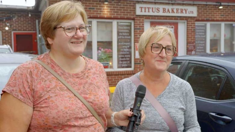 Two smiling women, both with short hair and wearing glasses, standing in front of Titus Bakery.