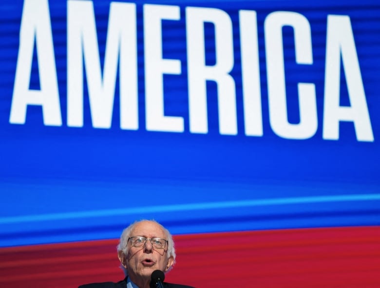 Bernie Sanders speaks on a stage at the Democratic National Convention, with the word America projected behind him.