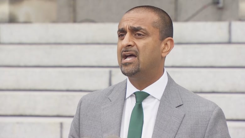 A man wearing a grey suit with a green tie standing outside behind grey steps. 