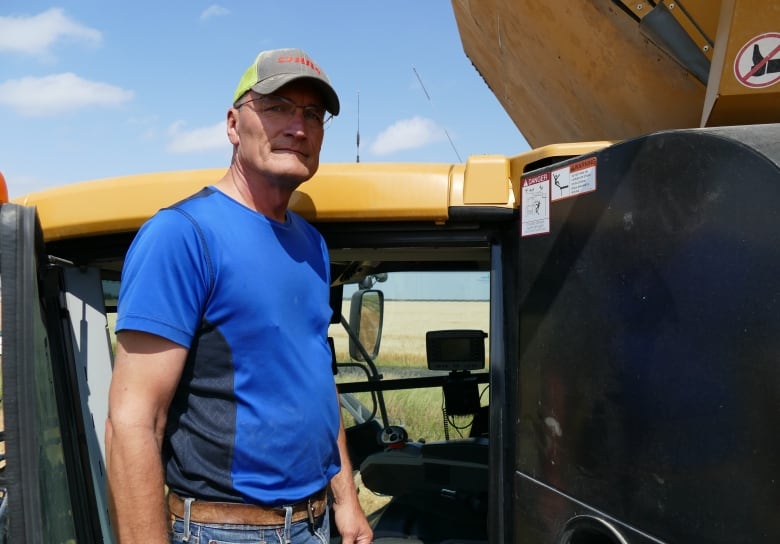 A man wearing a blue short and a ball cap stands on the step of his combine in a wheat field. 