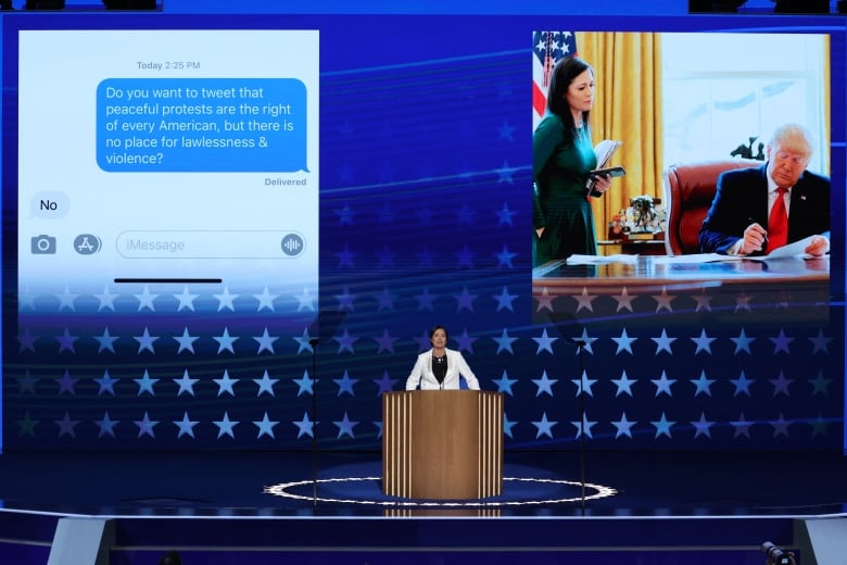 Long shot of woman on stage. In background a screen showing a picture of her in the Oval Office with Trump, and a picture of a text exchange with Melania