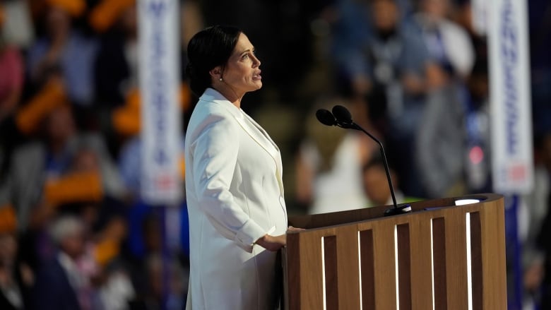 A brunette woman wearing a white blazer speaks at a podium.