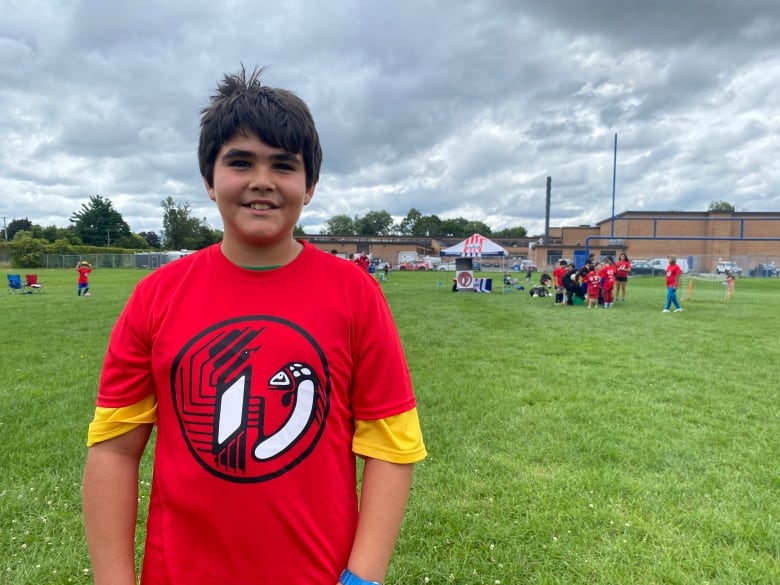 A child wearing the logo of the Odawa Native Friendship Centre stands on a field. A crowd of other children are off in the distance. 