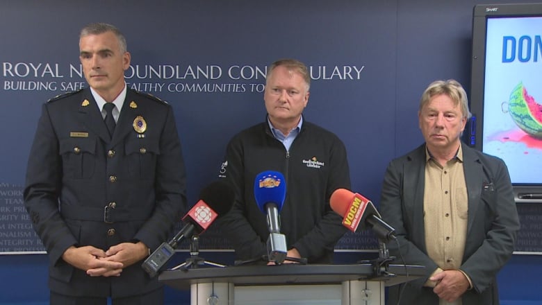 Three man stand behind a podium. The first wearing a police officer uniform, and the other wearing casual wear. 