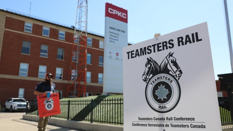 A man carrying a Teamsters Canada Rail Conference walks in the background of the photo while a white Teamsters Rail sign appears in foreground. 