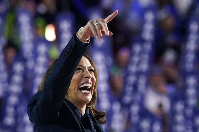 A woman in a dark suit laughs on a stadium stage and points. 