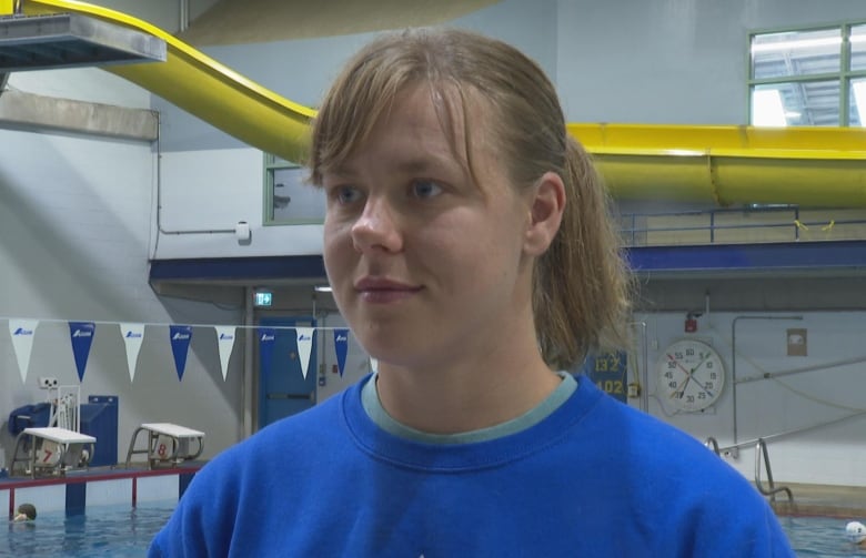 Woman, wearing blue sweater, standing in front of a swimming pool, looking at camera with a neutral expression.