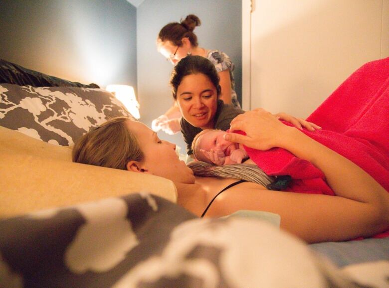 A newborn baby lies on its mother's chest as another woman looks over them.