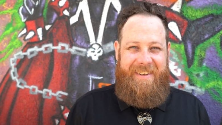 A brunette haired man with a red beard stands smiling in front of a wall of graffiti. He's wearing a black dress shirt and silver bolo tie.