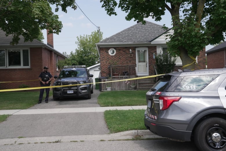 A police officer stands next to a police vehicle in a driveway of a brick house that's been taped off by police tape. A second police vehicle is parked on the street curb. It's a residential area. It's day time.