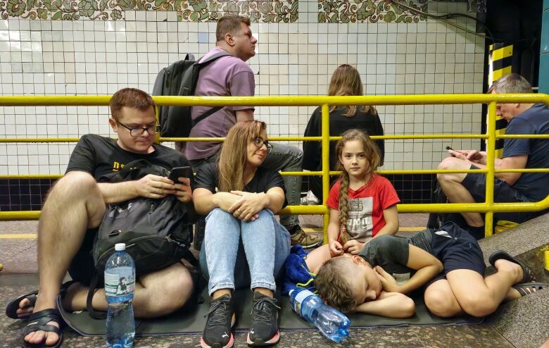 Adults and children are shown sitting and standing inside what appears to be a subway station. A boy lays on a mat near his family.