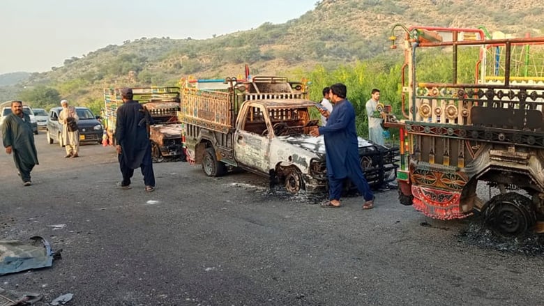 Burned out vehicles sit empty on a road in Pakistan, with a few men walking or standing nearby. 