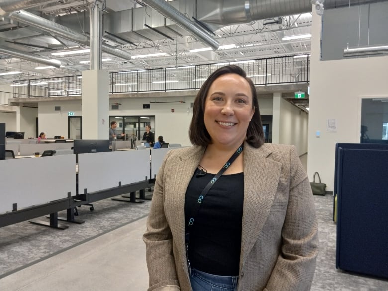 Woman in tweed blazer and black shirt smiling. In the background is a row of office desks