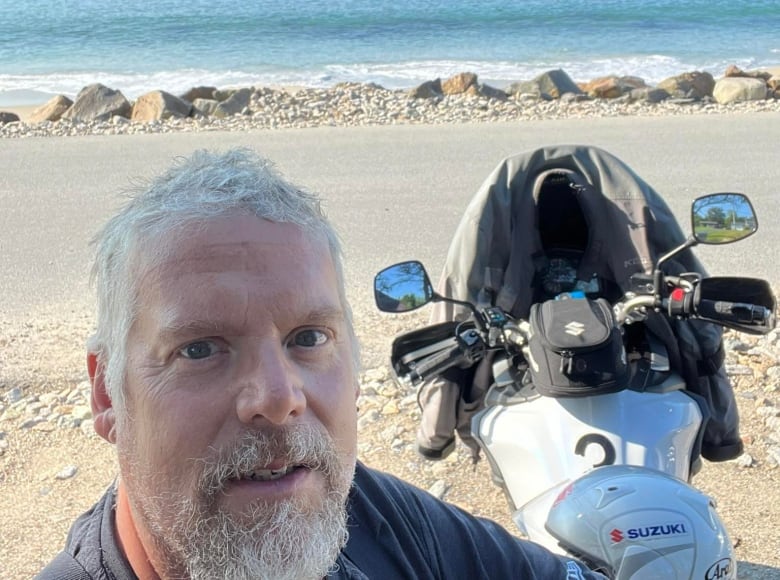 A man stands on a beach near a motorcyle.