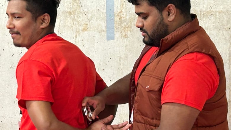 Two brown men wearing red t-shirts. One is putting handcuffs on the other during a police training workshop.