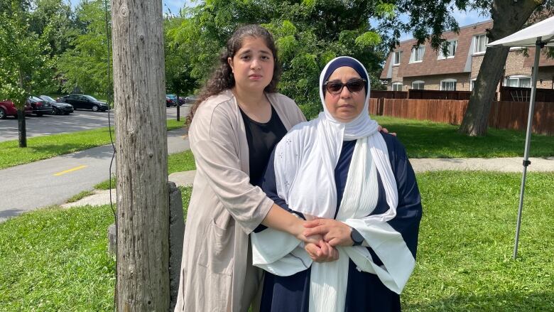 A young brunette woman dressed in beige wraps her arms around her mother, who us wearing a white hijab and sunglasses, as they pose outside their home.