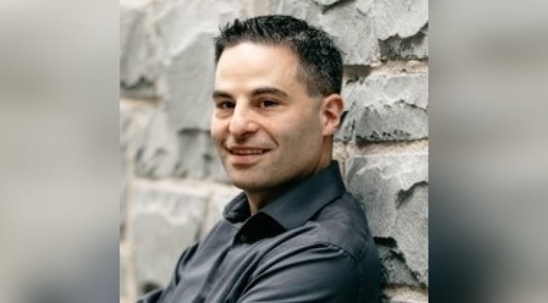 A man with gelled back silver hair and wearing a grey dress shirt smiles and leans against a stone wall.