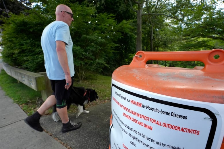 A passer-by walks a dog, Monday, Aug. 26, 2024, while entering a walkway, in Plymouth, Mass., near a sign that advises people of a ban in effect for outdoor activity between dusk and dawn due to the risk of exposure to mosquito-borne diseases.