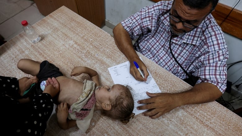 A man with glasses and a stethoscope sits at a desk writing on paper next to a toddler lying on her back, her belly exposed. 