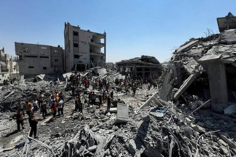 A crowd of people stand in the grey rubble of several buildings. A bombed out shell of a building is in the background. 