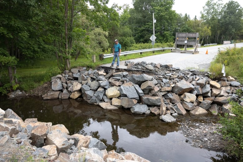 A man stands overlooking a large gap in the road where there was once a bridge over a stream. 