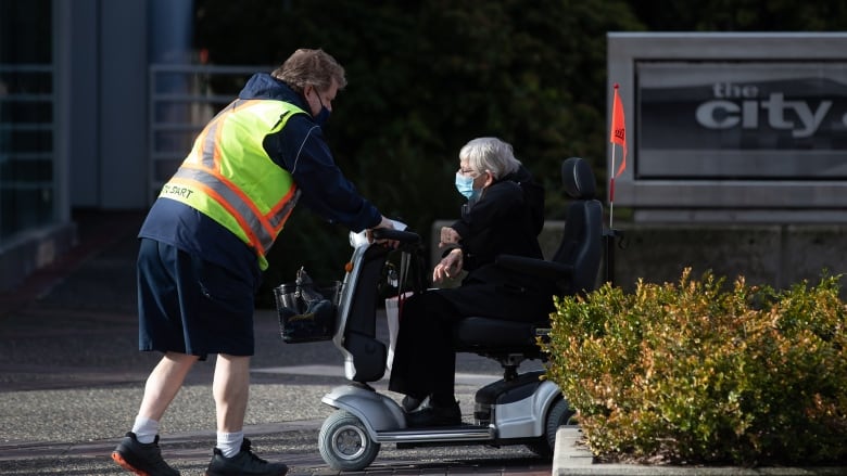 A man in a yellow vest helps an elderly woman on a mobility scooter.