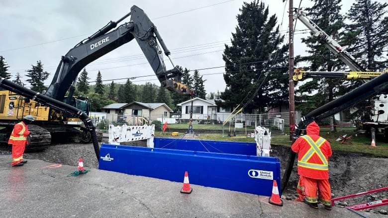 Construction workers stand around an open excavation site. It is raining.