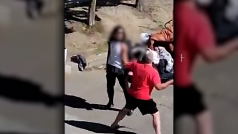 A grainy photo of a man in a red shirt standing and making a threatening gesture with a stick toward another man on the street outside the supportive housing building.