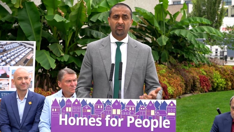 A man sporting a goatee and wearing a grey suit and green tie speaks in Kelowna from a podium that says Homes for People across it, as members of Kelowna city council sit behind him. 