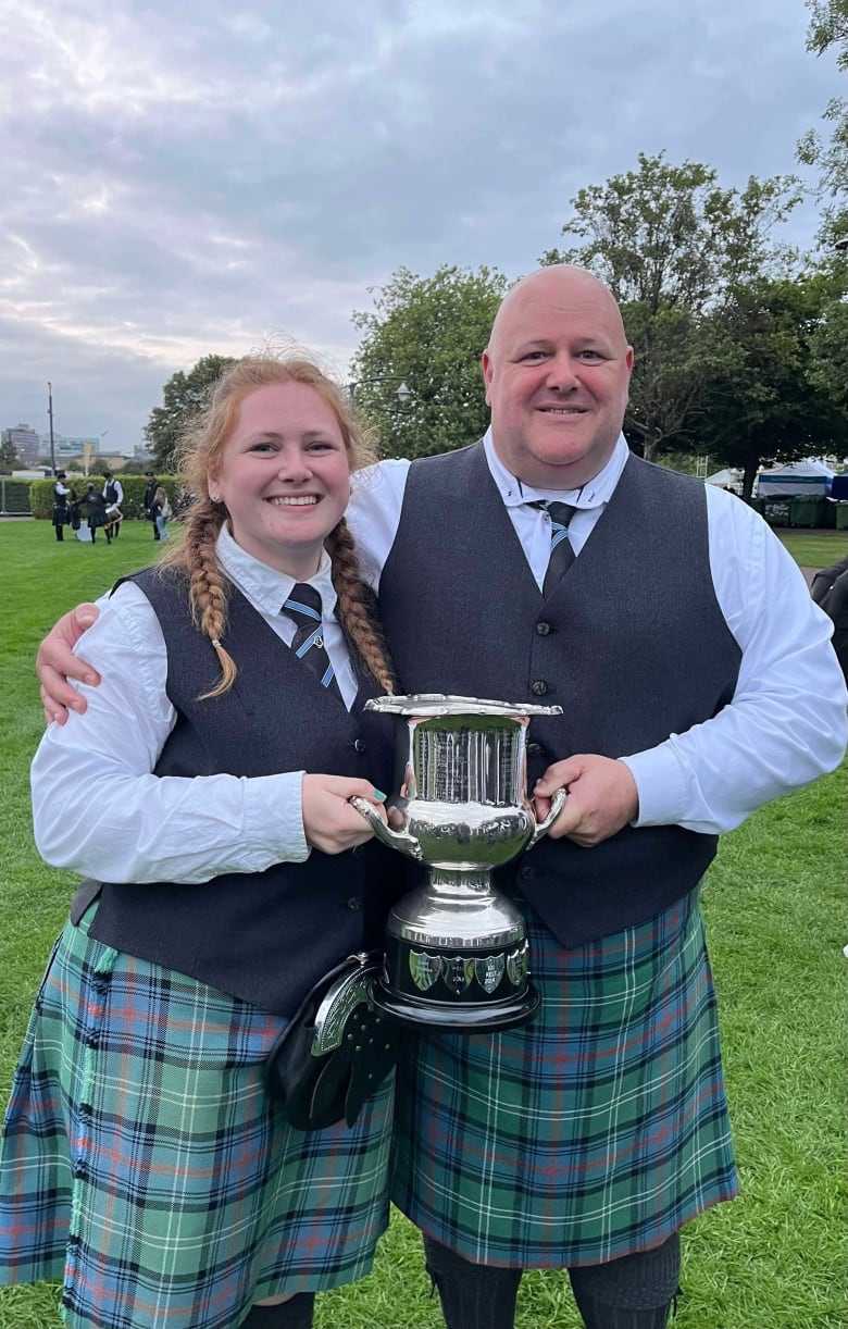 An young woman and her father, wearing Celtic attire including kilts, stand together holding a silver trophy.