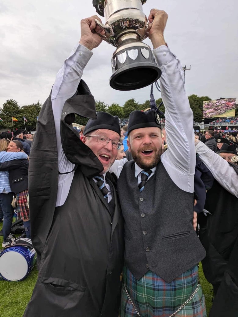 Two men wearing Scottish attire stand together and hold a silver trophy above their heads in celebration.