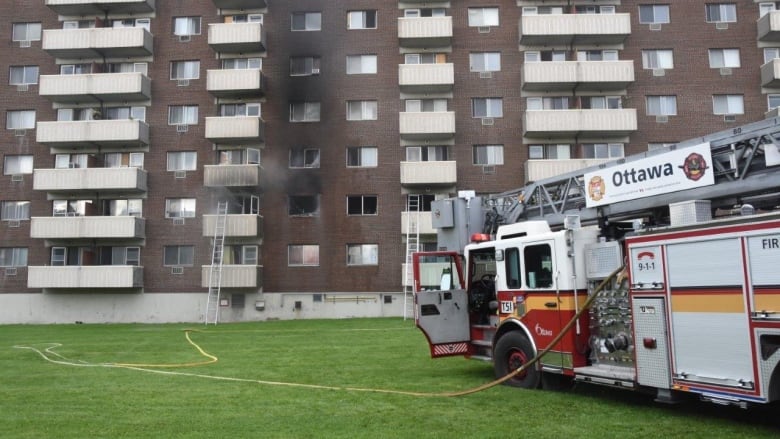 A fire truck outside a brick highrise building in summer. Hoses run from the truck to the building, which has light smoke coming from some open windows.