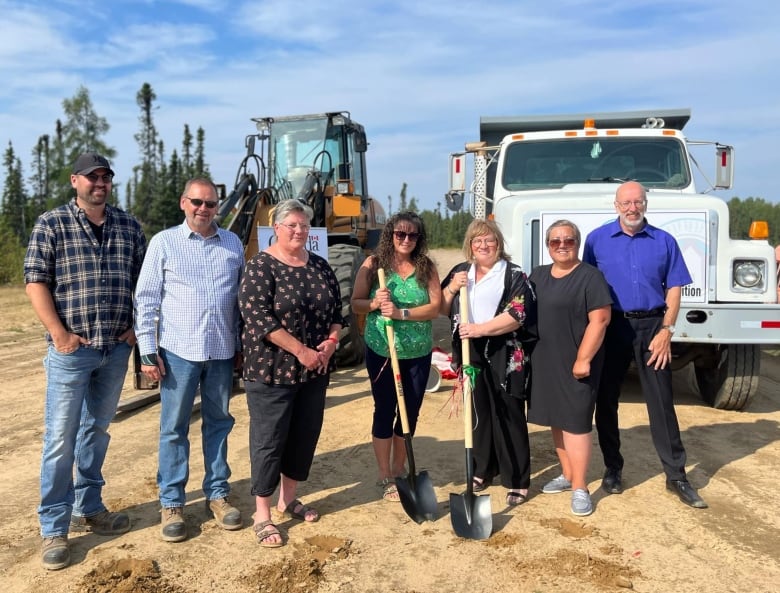 A group of seven people stand on a construction site. Two women in the front are holding shovels.