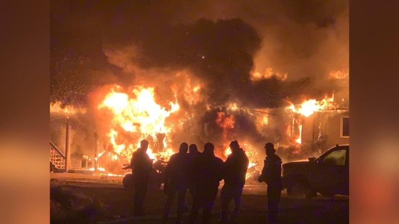 The silhouettes of six people are seen in front of a structure on fire.