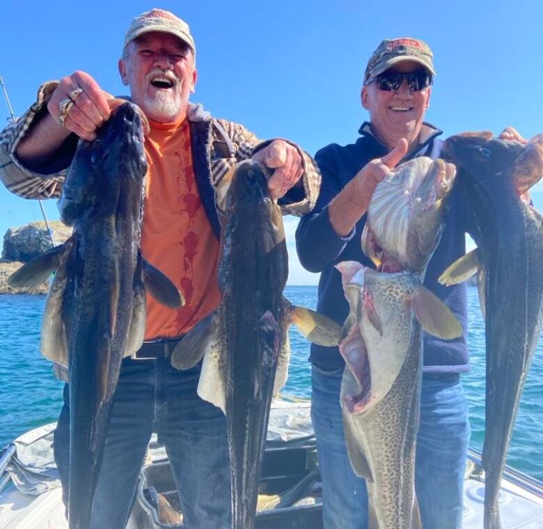 Two smiling men stand in a boat, holding up two fish each.