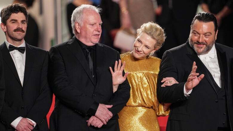 Three men in suits and a woman in a gold dress pose together.