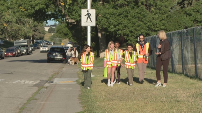 Four children wearing yellow reflective vests accompanied by an adult with an orange reflective vest and a woman wearing a black outfit.