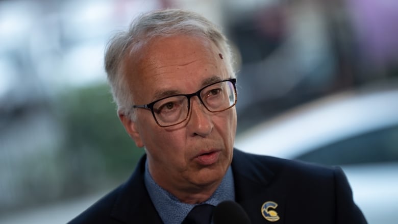 A white-haired man in glasses wearing a dark suit jacket over a dark blue shirt.