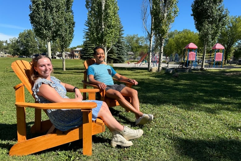 Two people sit in wooden chairs in a park under the blue sky with a playground behind them.