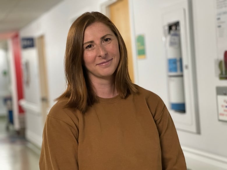 A woman with auburn hair stands in a hospital with her head titled to the side.